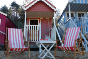 Beach hut with deck chairs and table set out in front. The beach hut is called "The Den" and is number 82 on Wells beach, Wells-next-the-Sea, North Norfolk. The hut is available to rent via Airbnb.