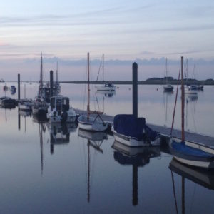 In the evening the wind often drops, making an evening tide in Wells-next-the-Sea extra beautiful. The skies and light of the magic hour are reflected on the water. The boats on the pontoons and moorings are also reflected in the still water. The East Hills can be seen in the background.